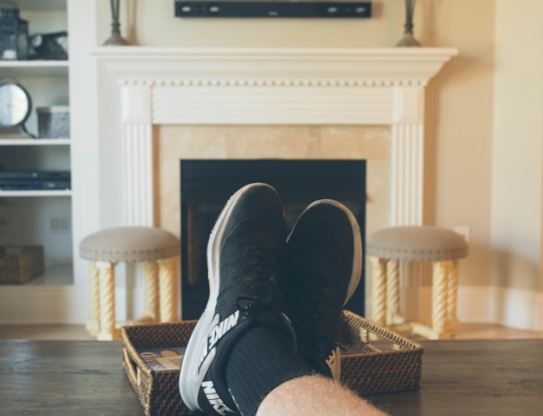 A person is relaxing in a living room with feet up on a wooden table. A TV mounted above a fireplace is displaying a popular streaming platform. Shelves on either side of the fireplace hold decorative items. A smartphone can be seen on the table.