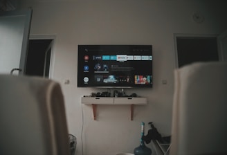 A living room setup featuring a wall-mounted TV displaying a streaming service interface. The room is dimly lit, with the TV as the main light source. Below the TV is a small shelf holding various objects. To the sides, there are partially visible chairs, and in the corner, there is a water bottle on the floor.
