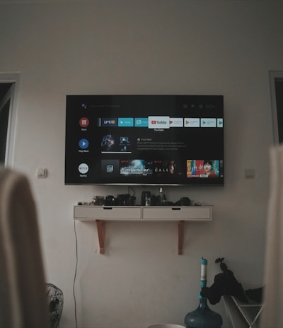 A living room setup featuring a wall-mounted TV displaying a streaming service interface. The room is dimly lit, with the TV as the main light source. Below the TV is a small shelf holding various objects. To the sides, there are partially visible chairs, and in the corner, there is a water bottle on the floor.