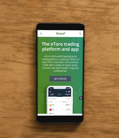 A smartphone displaying an advertisement for an online trading platform called eToro. The screen shows a green background with text promoting the platform as a leading social trading platform. A button labeled 'Get Started' is visible, along with a partial view of a mobile app screen below.