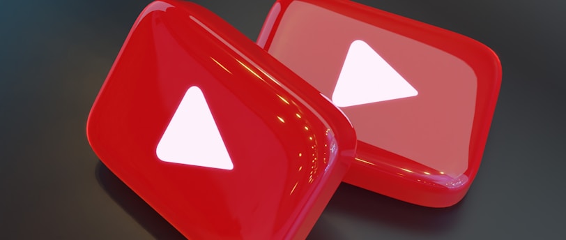 Two shiny, red, three-dimensional shapes resembling play buttons are positioned on a dark surface. Each button displays a white triangle in the center, which is characteristic of a popular video sharing platform.