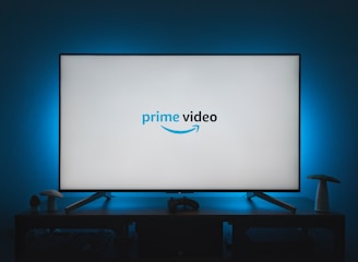 A television screen displays the Prime Video logo, situated on an entertainment unit in a dimly lit room. The room is illuminated with a subtle blue backlight, creating a calming atmosphere. Two small decorative items, resembling mushrooms, are placed on either side of the unit.