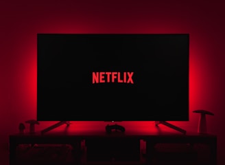 A dark room featuring a television screen displaying the word 'Netflix' in red against a black background. The screen is centered on a console table, with a video game controller placed in front. The room is dimly lit by red ambient lighting, creating a cozy atmosphere.