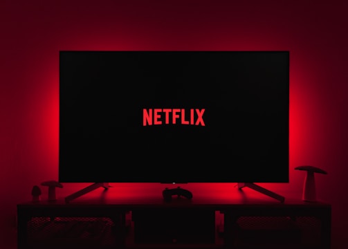 A dark room featuring a television screen displaying the word 'Netflix' in red against a black background. The screen is centered on a console table, with a video game controller placed in front. The room is dimly lit by red ambient lighting, creating a cozy atmosphere.