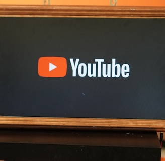 A rectangular screen encased in a wooden frame displaying the YouTube logo, characterized by a red play button icon next to the word 'YouTube' on a black background. The screen is placed on a small black table in a room with an orange wall and a couch in the background.
