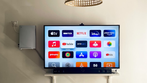 A modern living room features a wall-mounted flat-screen television displaying multiple streaming app icons, such as Apple TV, HBO Max, Netflix, Disney+, and Prime Video. Below the TV is a soundbar sitting on a small white shelf. A wicker pendant light hangs from the ceiling above, and the wall is painted a neutral color.