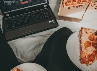 A cozy setting with a laptop displaying a streaming service interface nearby, alongside a pizza box holding a partially eaten pizza. Two slices of pizza are on paper plates held by someone, suggesting a casual movie night atmosphere.