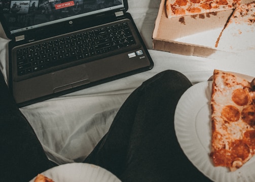 A cozy setting with a laptop displaying a streaming service interface nearby, alongside a pizza box holding a partially eaten pizza. Two slices of pizza are on paper plates held by someone, suggesting a casual movie night atmosphere.